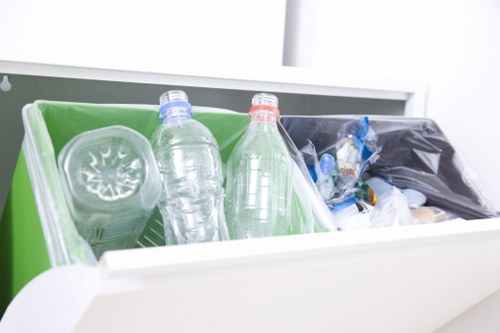 A close-up view of a white waste collection drawer or bin containing various types of rubbish, including a transparent plastic bottle with a blue cap, another similar bottle with a red cap, and a partially visible black plastic bag filled with miscellaneous waste. The interior of the drawer shows a bright green plastic tray on the left side, supporting the bottles, with the black bag situated on the right. The background reveals a plain white surface, likely part of a domestic or professional rubbish removal setup, placed in a clean environment that emphasizes the disposal of small plastic items typically managed by house clearance services in Baldock or surrounding areas.