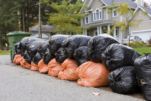 A row of tightly packed black and orange rubbish bags lined up along the edge of a residential street, with the black bags on top and the orange bags at the bottom, all filled with waste. Behind the bags, there are green wheelie bins positioned on the pavement, with some trees and a house with a light-colored exterior, multiple windows, and a gabled roof visible in the background. The scene takes place during daylight, and the bags appear to be prepared for rubbish removal services, reflecting typical urban or suburban waste collection practices near a property in Baldock or nearby areas. The image emphasizes the purpose of rubbish disposal in a residential environment, possibly by a professional rubbish clearance company like House Clearance Baldock.