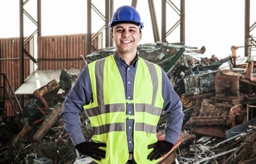A male worker dressed in a blue shirt, high-visibility yellow vest, black gloves, and a blue safety helmet poses inside an industrial warehouse or scrapyard with a smiling expression. Behind him, there is a large pile of assorted metal scraps, car parts, and debris, suggesting a site for rubbish or scrap removal. The background features metal framework and large windows, allowing natural light to illuminate the scene. The worker's stance and the environment indicate a professional setting related to rubbish clearance or waste management services in Baldock, reflecting the activities managed by House Clearance Baldock in the local area. The image emphasizes safety equipment and a workspace dedicated to clearing and managing industrial or household waste materials.