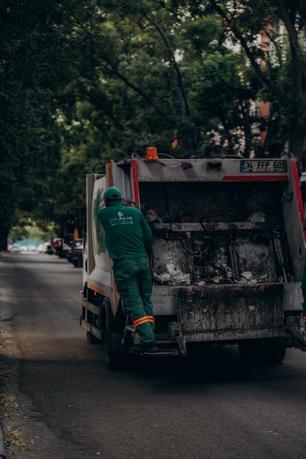 A sanitation worker in a green uniform with orange reflective stripes is standing beside the rear of a large waste collection vehicle on a quiet urban street lined with trees and parked cars. The truc