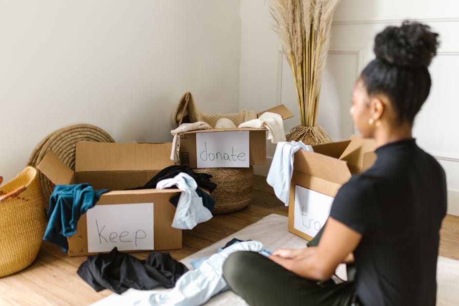 A woman with dark, curly hair tied in a high bun, dressed in a black top, is seated on a light-colored wooden floor next to a collection of open cardboard boxes filled with used clothing. Each box is 