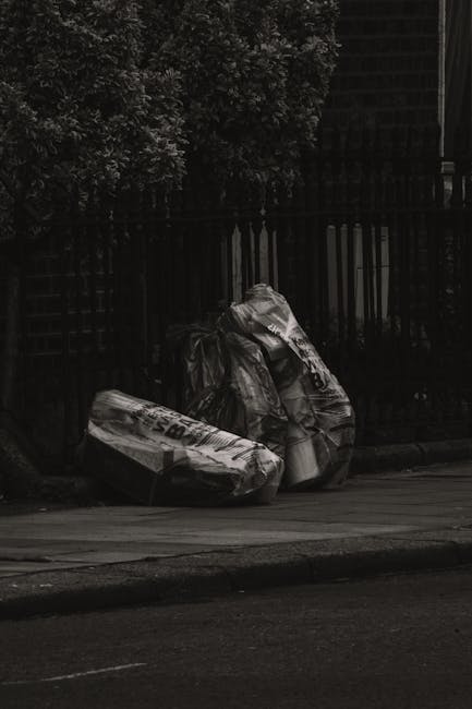 A pile of mixed waste materials, including cardboard boxes, paper bags, and plastic packaging, is gathered in the corner of an outdoor area adjacent to a brick and concrete wall. The cardboard boxes a